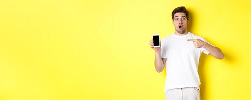 Impressed guy check out smartphone screen, pointing finger at display and gasping amazed, standing over yellow background photo