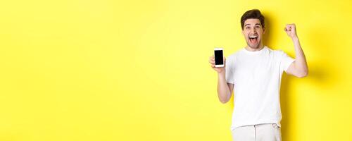 Cheerful guy showing smartphone screen, raising hand up and celebrating, triumphing over internet achievement, standing over yellow background photo