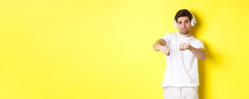 Cool guy listening music in headphones and dancing, standing in white clothes against yellow studio background photo