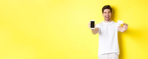 Happy man showing good online offer on mobile phone screen, holding credit card and winking, standing over yellow background photo