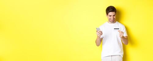 Guy making online order, register credit card in mobile application, holding smartphone and smiling, standing over yellow background photo