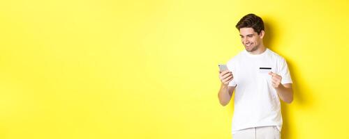Young man paying online, insert credit card number on mobile phone, shopping in internet, standing over yellow background photo