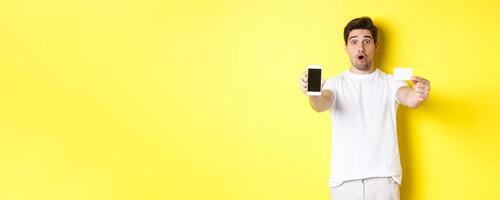 Handsome caucasian guy showing smartphone screen and credit card, concept of mobile banking and online shopping, yellow background photo