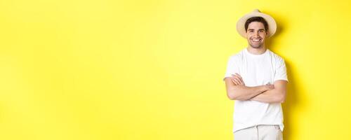 Tourism. Handsome young man looking happy, wearing straw hat for travelling, cross hands on chest and smiling, standing over yellow background photo