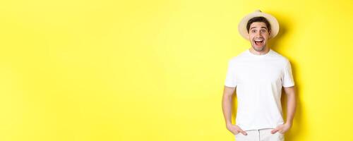 Surprised man tourist in straw hat looking happy, react amazed to travel agency advertisement, standing over yellow background photo