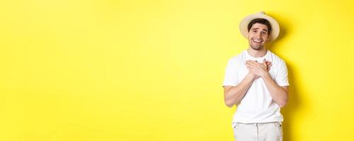 Concept of tourism and summer. Grateful guy in straw hat holding hands on heart, saying thank you and smiling with gratitude, standing against yellow background photo