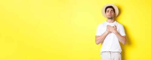 Concept of tourism and summer. Romantic man in straw hat looking nostalgic, close eyes and holding hands on heart, standing against yellow background photo