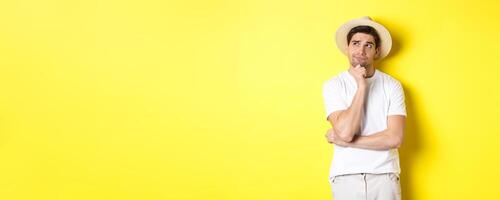 Concept of tourism and summer. Thoughtful man tourist pondering, looking at upper left corner and thinking, standing in straw hat and white t-shirt against yellow background photo