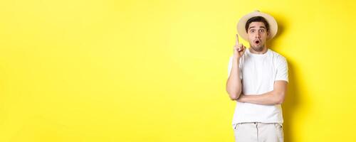 Portrait of young man in straw hat having an idea, raising finger eureka sign, making suggestion, standing over yellow background photo