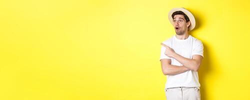 Concept of tourism and lifestyle. Excited handsome guy in straw hat checking out advertisement, pointing and looking at upper left corner logo, yellow background photo