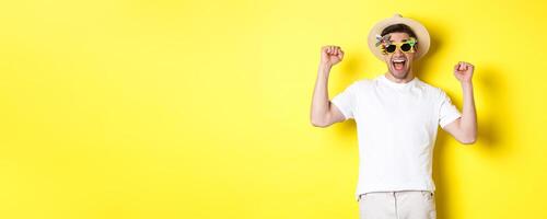 Concept of tourism and lifestyle. Happy man winning trip to resort, shouting yes and raising hands up, triumphing, wearing sunglasses and summer hat, yellow background photo