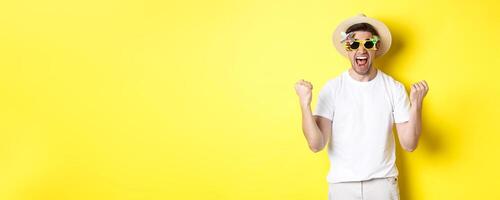 Happy guy going on vacation, winning or celebrating, wearing summer hat and sunglasses. Tourist looking excited, standing against yellow background photo