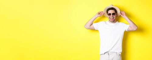 Happy man on vacation, wearing straw hat and sunglasses, smiling while standing against yellow background photo