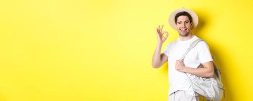 Tourism, travelling and holidays concept. Happy tourist going on vacation, holding backpack and showing okay sign smiling satisfied, standing against yellow background photo