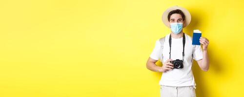 Concept of covid-19, travelling and quarantine. Happy man tourist with camera, showing passport and tickets for vacation, going on trip during pandemic, yellow background photo