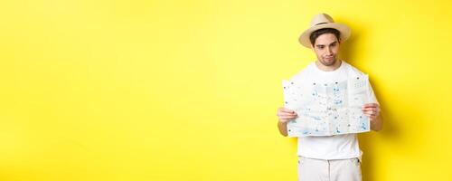 Travelling, vacation and tourism concept. Male tourist looking confused at map, looking for right direction, standing over yellow background photo
