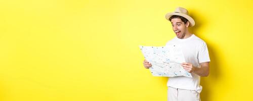 Travelling, vacation and tourism concept. Smiling happy tourist looking at map with sightseeings, standing against yellow background photo