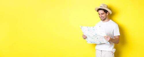 Travelling, vacation and tourism concept. Smiling happy tourist looking at map with sightseeings, standing against yellow background photo
