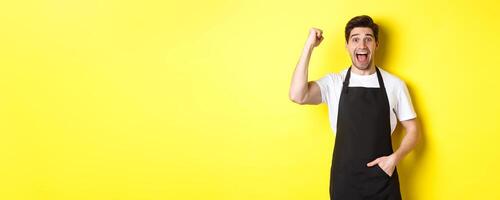 Cheerful seller making fist pump, rejoicing and triumphing, standing in black apron against yellow background photo