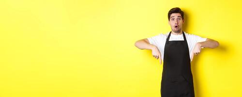 Surprised seller in black apron pointing fingers down, looking amazed at camera, standing over yellow background photo