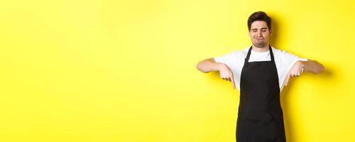 Doubtful barista complaining, pointing fingers down and grimacing displeased, standing in black apron against yellow background photo