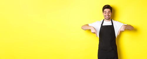 Happy coffee shop worker in black apron pointing fingers down, looking pleased and smiling, showing logo, standing over yellow background photo