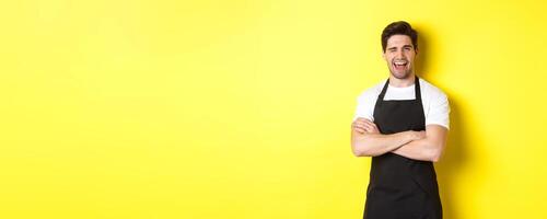 Handsome barista in black apron winking at you, wearing black apron uniform, standing over yellow background photo