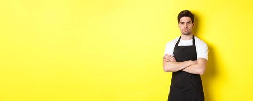 Suspicious barista squinting, cross arms on chest and looking at something with disbelief, standing over yellow background photo