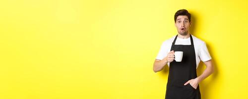 Barista holding coffee mug and looking surprised, standing in black apron cafe uniform against yellow background photo