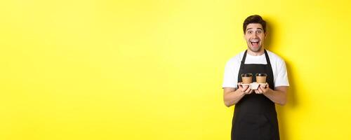 Excited male barista holding two cups of takeaway coffee, working in cafe, standing over yellow background photo