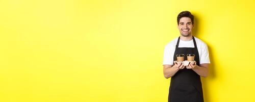 Handsome male barista serving takeaway coffee and smiling, bringing order, standing in black apron against yellow background photo