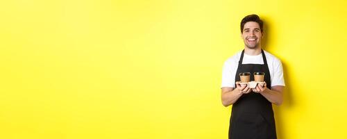 Friendly barista in black apron giving takeaway order, holding two cups of coffee and smiling, standing over yellow background photo