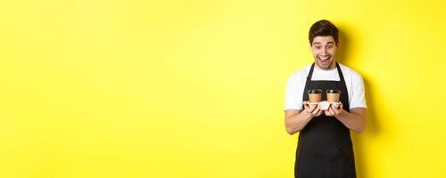 Waiter looking excited at two cups of takeaway coffee, wearing black apron, standing over yellow background photo