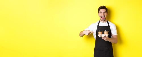 Excited barista in black apron pointing fingers at takeaway coffee cups, standing against yellow background happy photo