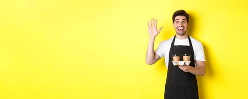 Friendly waiter in cafe waving hand at customer, holding takeaway coffee oder, standing against yellow background in black apron photo