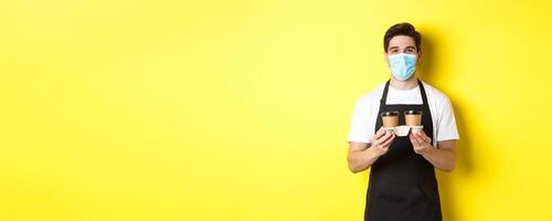 Concept of covid-19, cafe and social distancing. Barista in medical mask serving coffee in takeaway cups, standing in black apron against yellow background photo