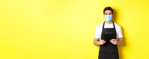 Concept of covid-19, small business and quarantine. Salesman in black apron and medical mask holding clipboard, working in shop, standing over yellow background photo