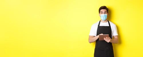Concept of covid-19, small business and pandemic. Waiter in black apron and medical mask taking order, holding digital tablet, standing over yellow background photo