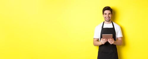 Waiter in black apron taking orders, holding digital tablet and smiling friendly, standing over yellow background photo