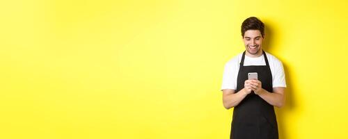 Waiter in black apron reading message on mobile phone, smiling happy, standing over yellow background photo