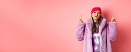 Stylish middle aged asian woman in purple fake fur coat, looking skeptical and grimacing with displeased face, pointing fingers up at something bad and unimpressive, pink background photo