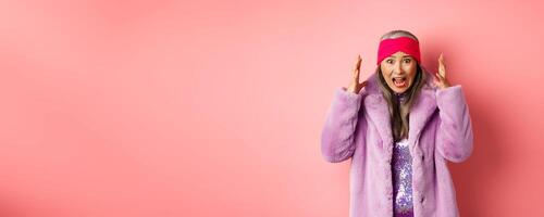 Anxious asian senior woman in stylish faux fur coat screaming, shaking hands and shouting alarmed and worried at camera, standing over pink background photo