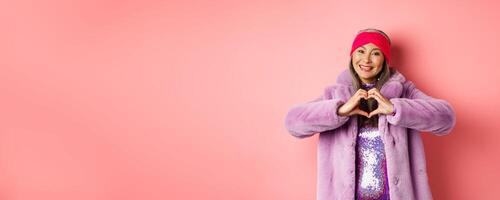 Romance and valentines day. Happy asian senior woman showing heart sign, I love you gesture, smiling and looking caring at camera, standing in faux fur coat, pink background photo