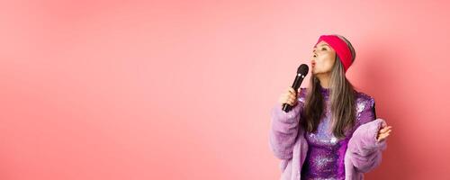 Gorgeous asian senior woman singing karaoke in mic, perform song and looking happy, standing over pink background photo