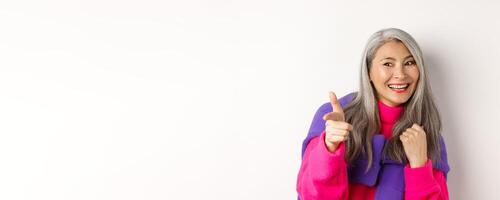 Close-up of happy and cool senior hipster woman, checking out, pointing and looking at person, smiling sideways, standing over white background photo