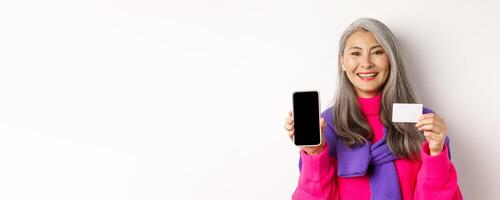 Online shopping. Closeup of smiling asian senior woman showing blank mobile screen and plastic credit card, looking happy, standing over white background photo