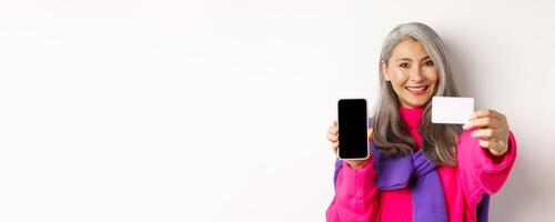 Online shopping. Close-up of stylish korean senior woman showing empty mobile screen and plastic credit card, smiling happy, standing over white background photo