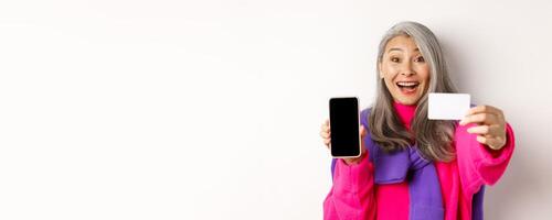 Online shopping. Close-up of stylish korean senior woman showing empty mobile screen and plastic credit card, smiling happy, standing over white background photo