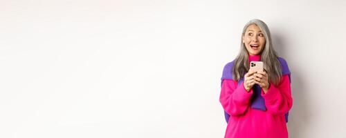 Amazed asian woman smiling and looking aside fascinated after reading promotion on smartphone, standing with mobile phone over white background photo