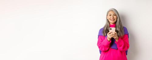 Happy asian woman smiling at camera after reading promotion on smartphone, standing with mobile phone over white background photo